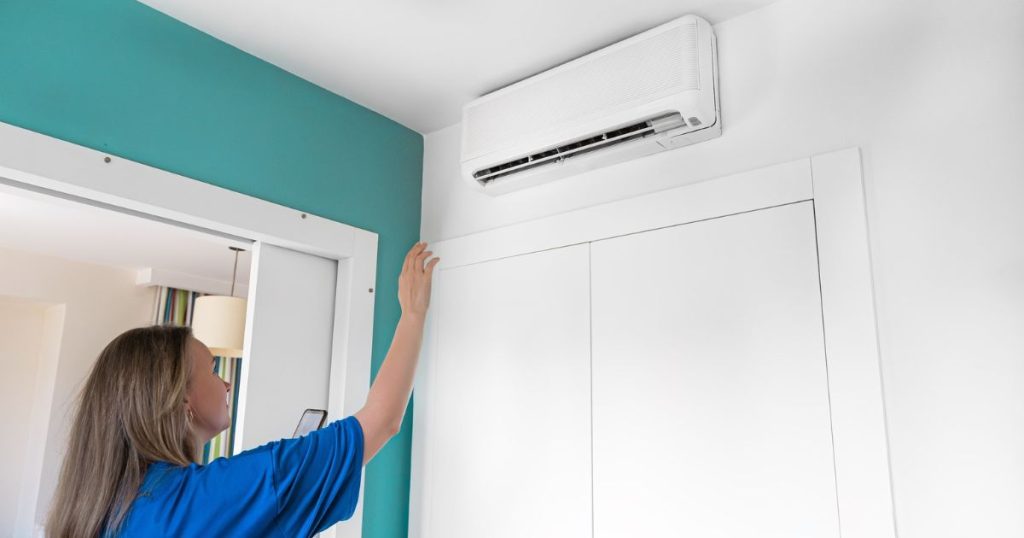 Een airco zonder buitenunit bevestigd aan een witte muur in een woning. Voor de airco staat een vrouw met een blauw shirt dat haar telefoon in een hand vast heeft en met haar andere hand omhoog staat om de lucht van de airco te voelen.