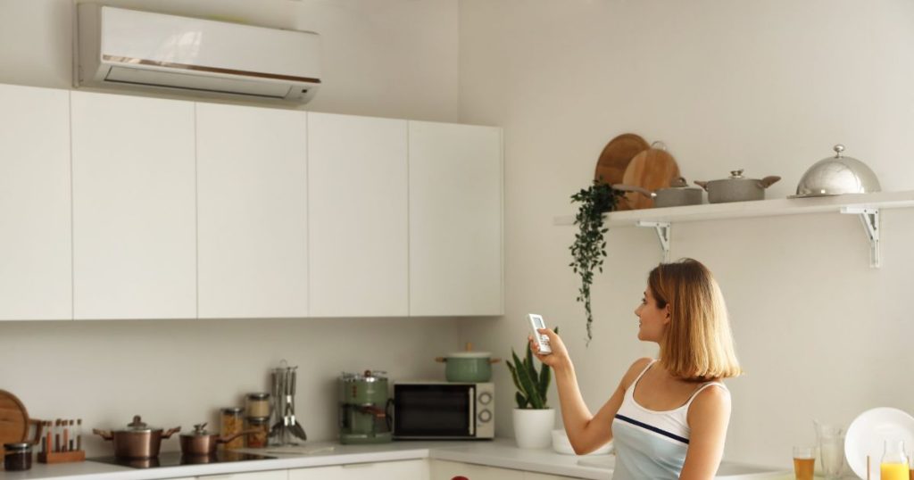 Een binnenunit airco in de keuken van een woning. De airco is boven een aantal witte keukenkastjes geplaatst. In de keuken staat een vrouw met een afstandsbediening waarmee ze de airco bedient.