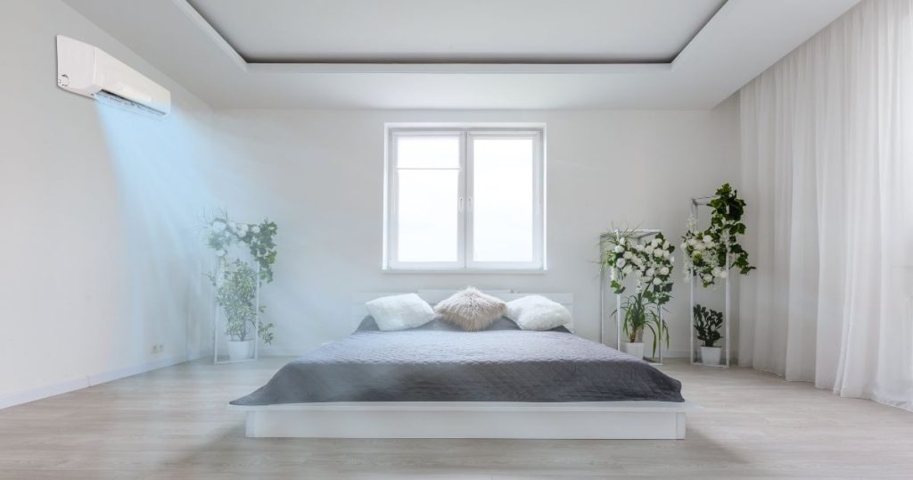 De binnenunit van een airco verspreidt koele lucht in een slaapkamer. In de slaapkamer is ook een groot bed te zien met een grijs dekbedovertrek. Naast het bed staan nog enkele kamerplanten en een wit gordijn hangt aan de muur.