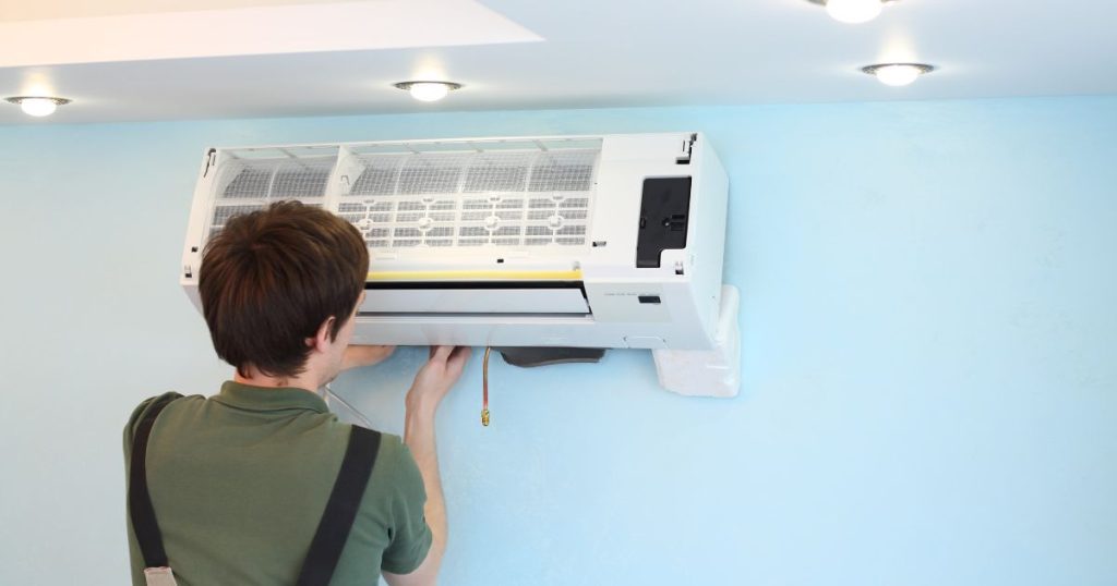 Jonge specialist plaatst de binnenunit van een airco tegen een blauwe muur. De airco is nog gedeeltelijk open.