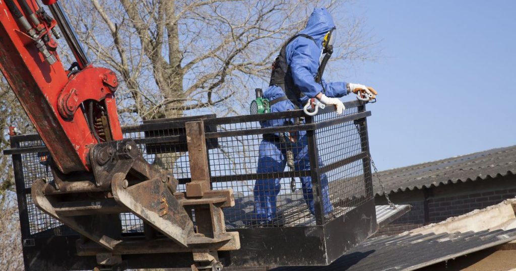 Een asbestspecialist in een blauw beschermingspak verwijdert een asbestdak.  