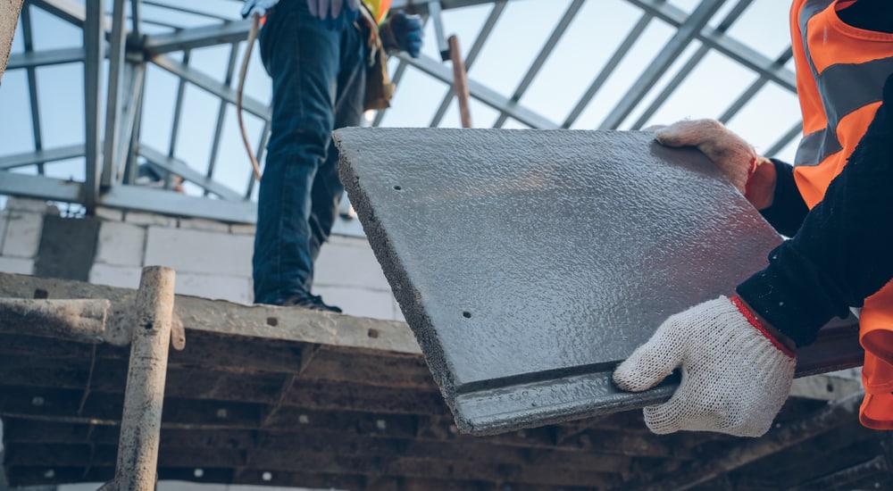 Een dakwerker met fluohesje en werkhandschoenen houdt een betonnen dakpan vast: een soort dakbedekking met een levensduur van 30 tot 50 jaar.