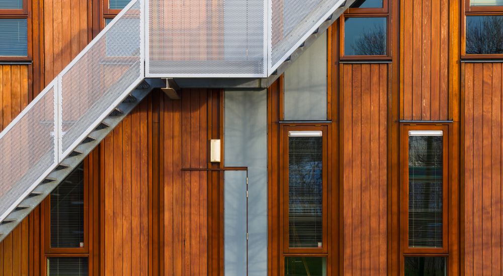 Een woning met houten gevelbekleding in verticale planken. De gevel heeft verschillende ramen. Langs de buitenmuur loopt een metalen trap die de verschillende verdiepingen van het gebouw met elkaar verbindt.