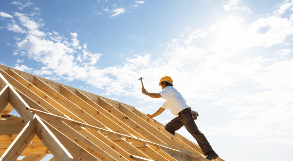 Een dakwerker met gele helm legt met zijn hamer de laatste hand aan de structuur van een gordingendak.