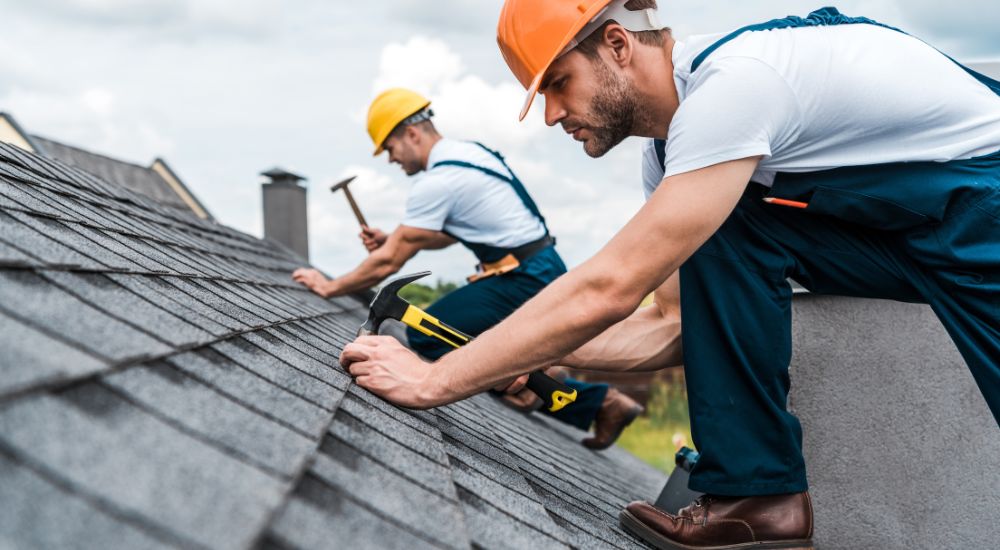 2 dakwerkers in blauwe overall en met een veiligheidshelm (geel en oranje) zijn druk in de weer met het vastnagelen van de donkere leien.