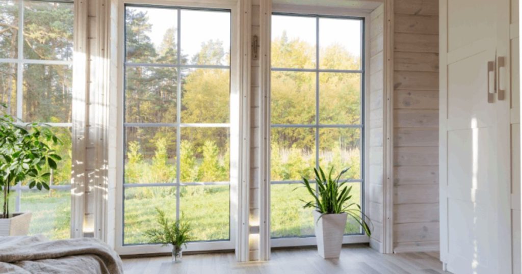 Grote witte houten ramen in een slaapkamer met uitzicht op de tuin die wordt omzoomd door lage struiken. Op de achtergrond is een bos zichtbaar.