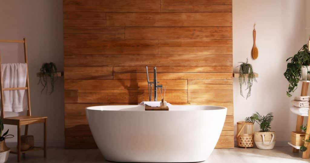 Een landelijke badkamer met een houten wand en veel planten met een losstaand bad. 