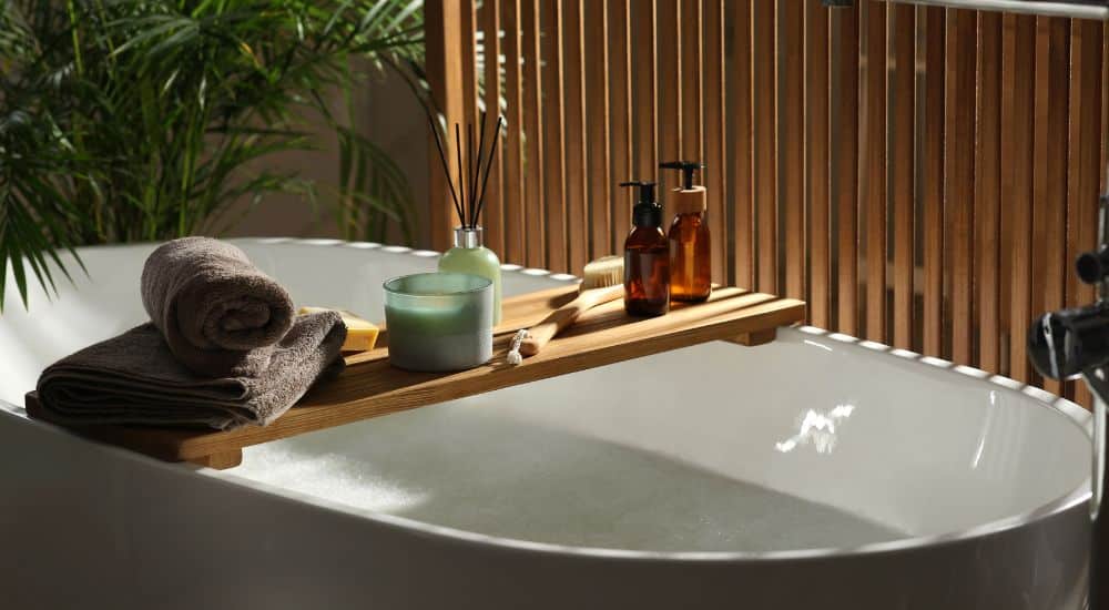 Een ruim wit losstaand ligbad met accessoires in een ruimte met houten accenten en een groene plant. 