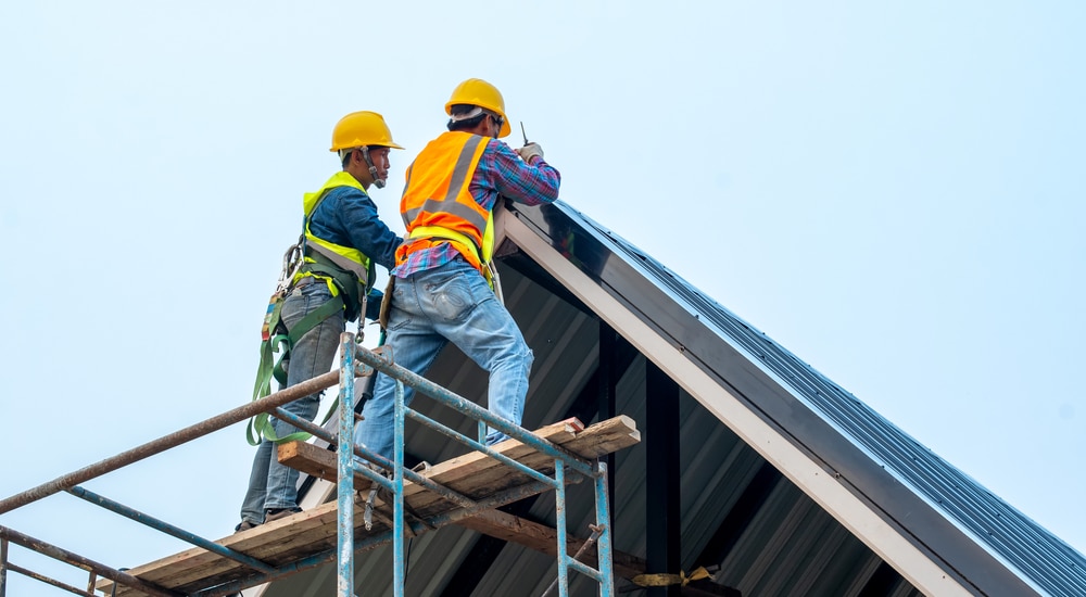 2 Dakwerkers met fluohesje, gele helm en veiligheidsriemen werken vanop een stelling aan de daknok van een aluminium dak.