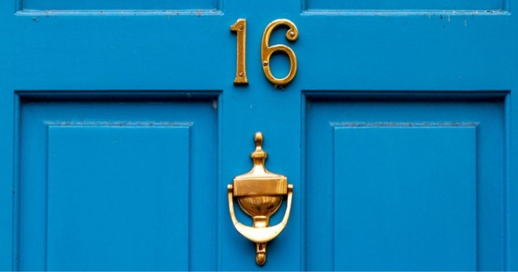 Een blauwe houten voordeur met huisnummer 16 en een hendel om aan te kloppen in koper.
