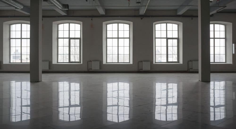 Een grote hal met grote stalen ramen. 2 ramen zijn voorzien van een insectengaas. Tussen elk raam hangt een chauffage aan de muur. Het kale interieur voelt kil en toch stijlvol aan. De ideale plek voor recepties en/of feesten?