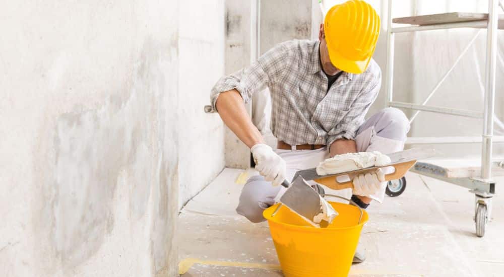 Een vakman met een gele helm op repareert een beschadigde muur met vochtproblemen.