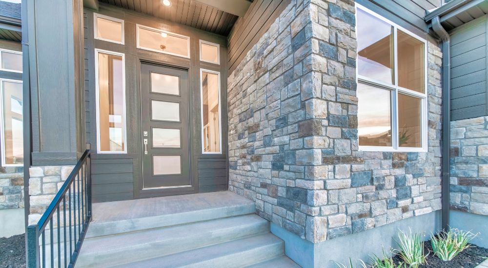 Een gevel met gesmoorde baksteen in verschillende formaten. De gevel steekt uit langs een trap die naar de voordeur leidt. De voordeur is omkaderd met ramen met witte raamkozijnen en houten gevelbekleding.