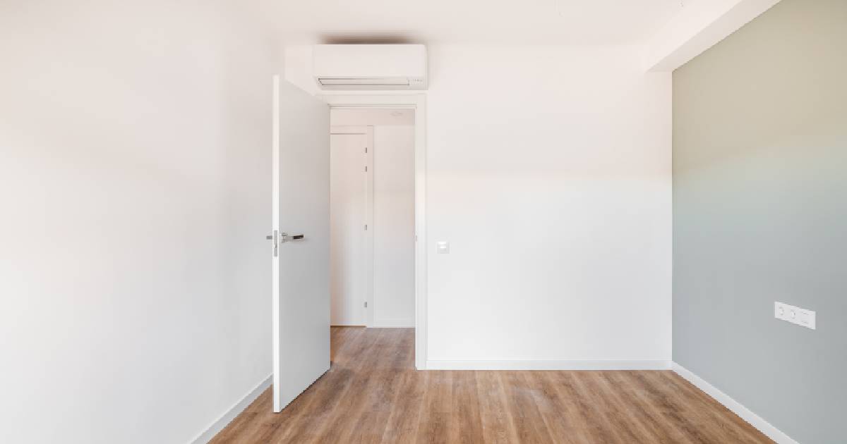 Een subtiele witte airco boven een deur in een witte kamer met houten vloer en grijsgroene accentmuur.
