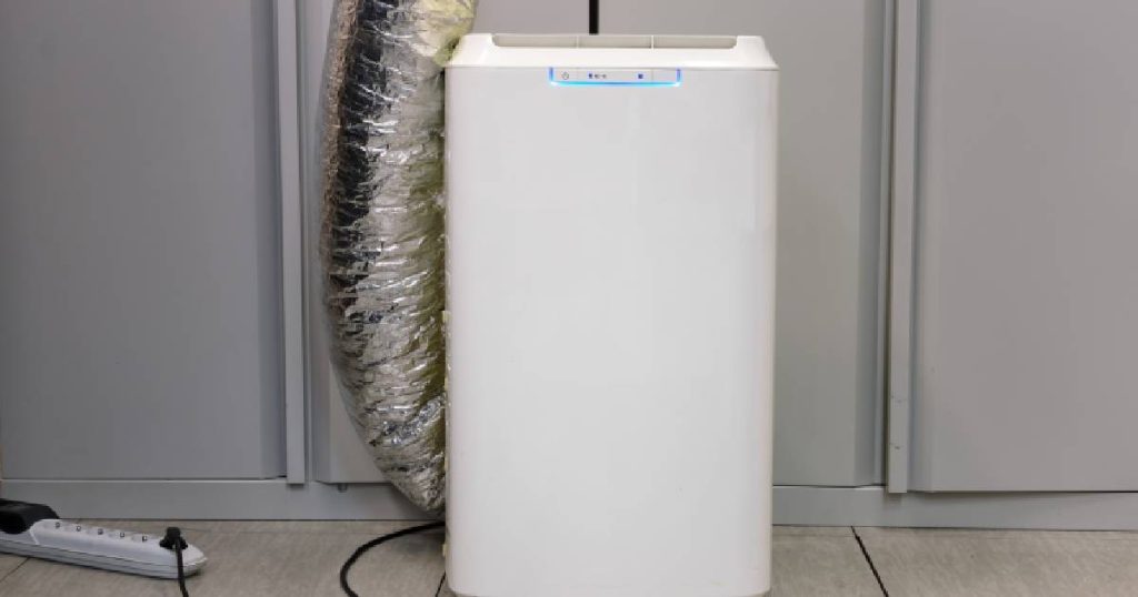 Een witte, compacte, verplaatsbare en draagbare mobiele airco op een tegelvloer. Een zilveren afvoerbuis leidt de oude, warme lucht naar buiten. Het toestel is aangesloten op een stopcontact met een eenvoudige stekker.