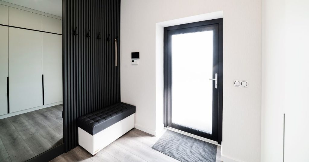 Een donkere aluminium voordeur met melkglas gefotografeerd langs de binnenzijde van de woning. Aan deur ligt een grijze mat op de grond.