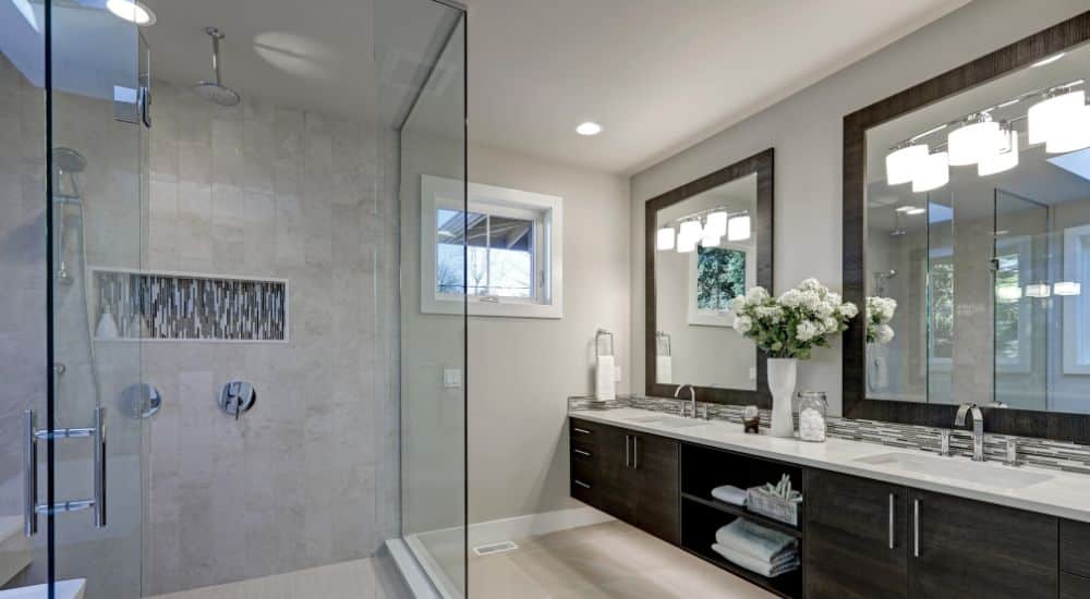 Een ruime badkamer met grijze muren en tegelvloeren, de douche heeft glazen wanden, grote spiegels boven de wastafels om de ruimte optisch te vergroten.