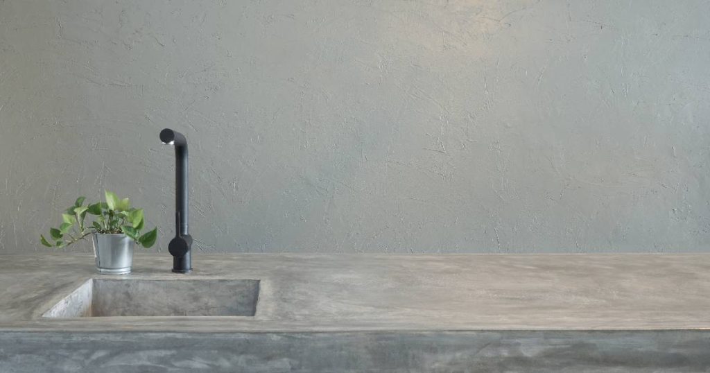Een keuken in betonlook met betonnen aanrecht en ingebouwde spoelbak met zwarte kraan, afgewerkt met een plantje in een metalen plantenpot.