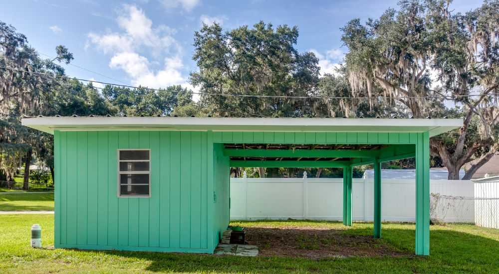Een tuinveranda met aangrenzend overdekt terras in felgroen hout met een wit dak tegen de witte omheining van een groene tuin.