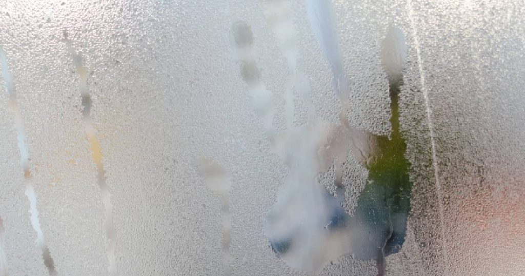 Condens op de binnenkant van een raam in een serre. In de condens is een handafdruk zichtbaar.