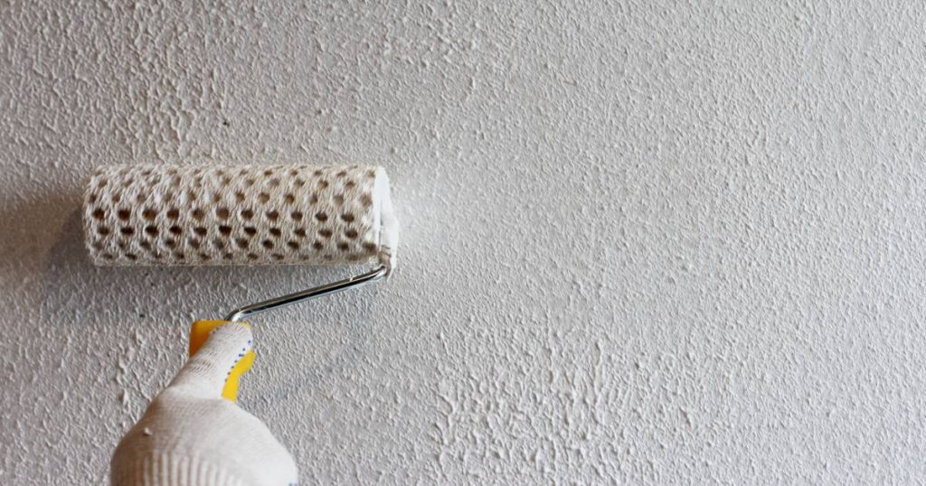 Witte sierpleister wordt met een roller op een binnenmuur aangebracht. De schilder draagt een witte handschoen.