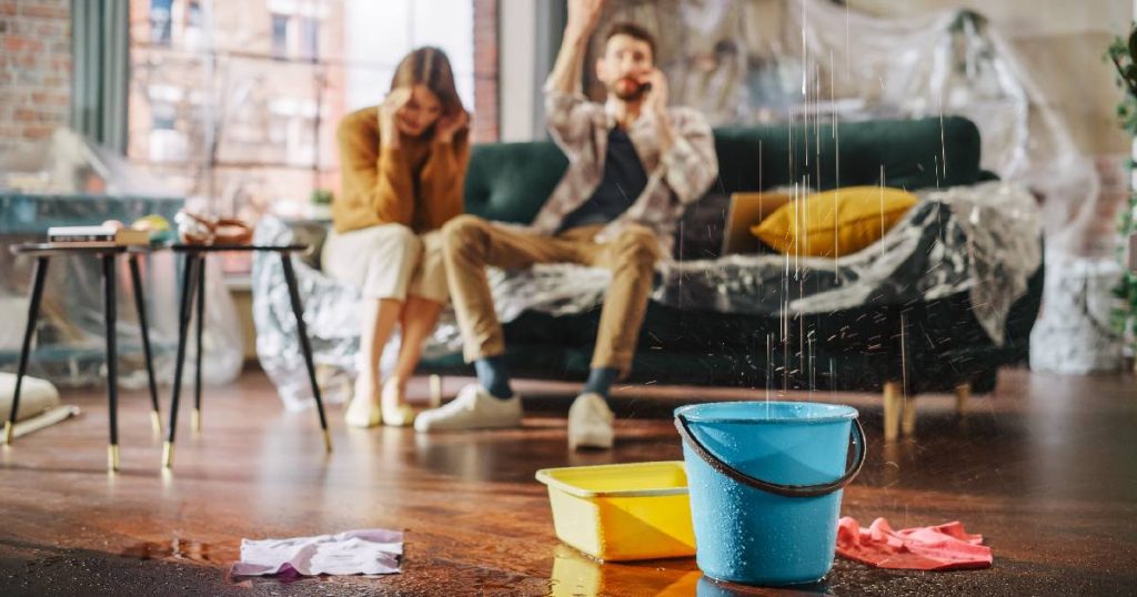 Een lichtblauwe emmer en geel teiltje vangen regendruppels op die binnen druipen door een daklekkage. Op de achtergrond een man en een vrouw op een bank: de man belt een dakdekker.