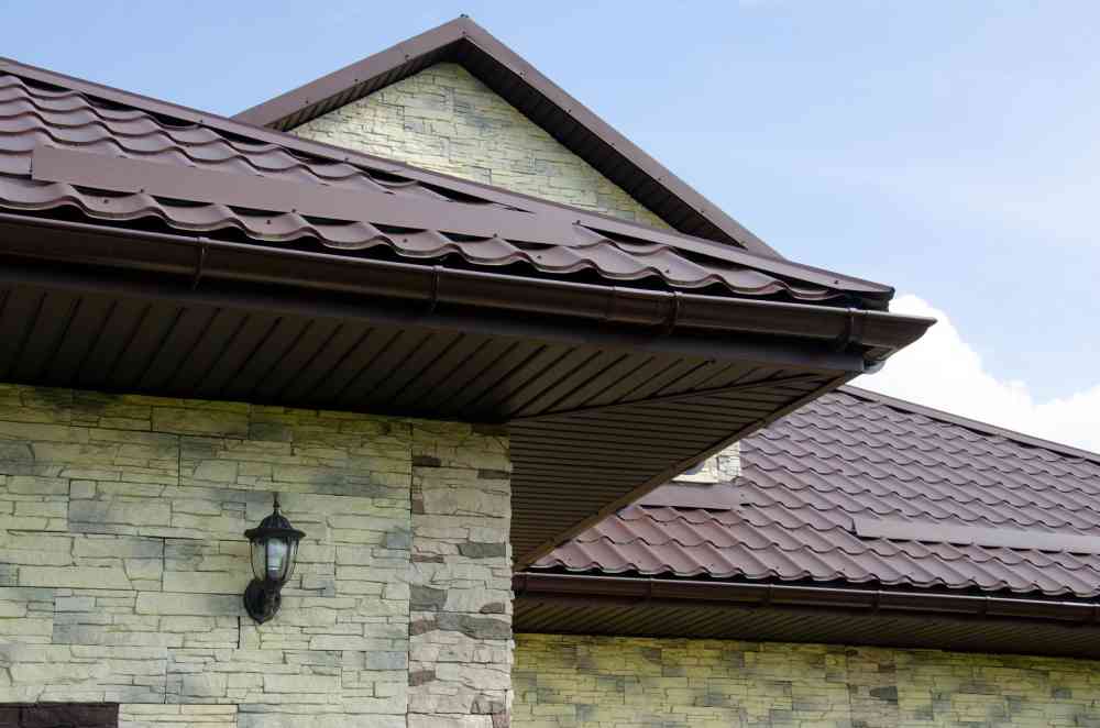 Een dakoverstek bij een woning met grijze dakpannen. De dakoverstek is onder de dakgoot geplaatst en ook in de bruine kleur uitgevoerd.
