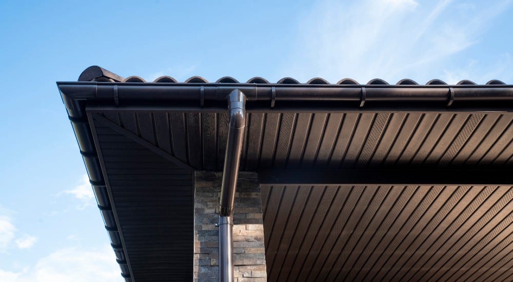 Een dakoverstek aan de onderkant van een dak met dakpannen. Ook is er een donkere dakgoot te zien met een regenpijn eronder.