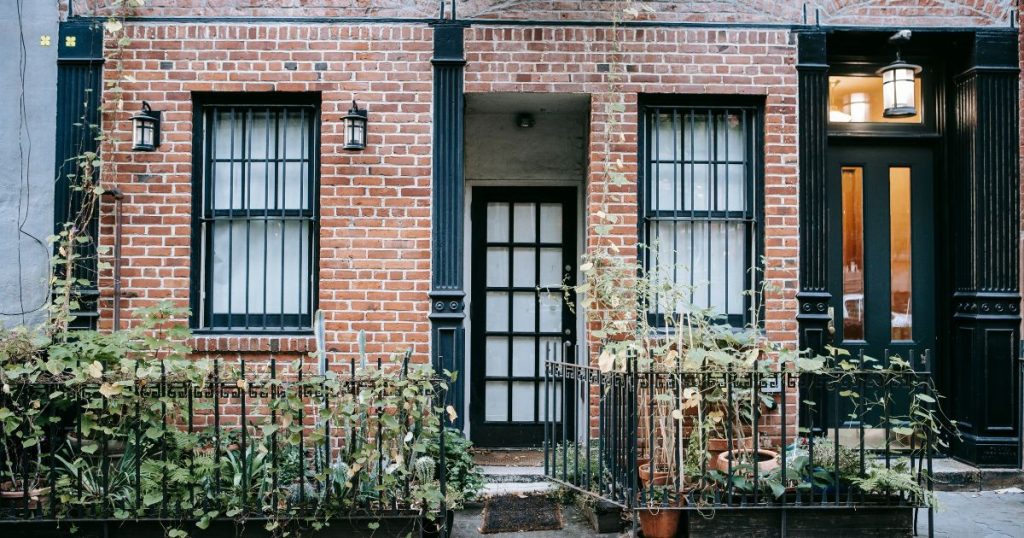 Een kleine gezellige woning met bakstenen gevel, zwarte raamkozijnen met zwarte kruishouten, en een voortuintje met smeedijzeren hekken en verschillende planten in terracotta potten. Naast het linkse raam hangen, langs weerszijden, kleine zwarte wandlantaarns.
