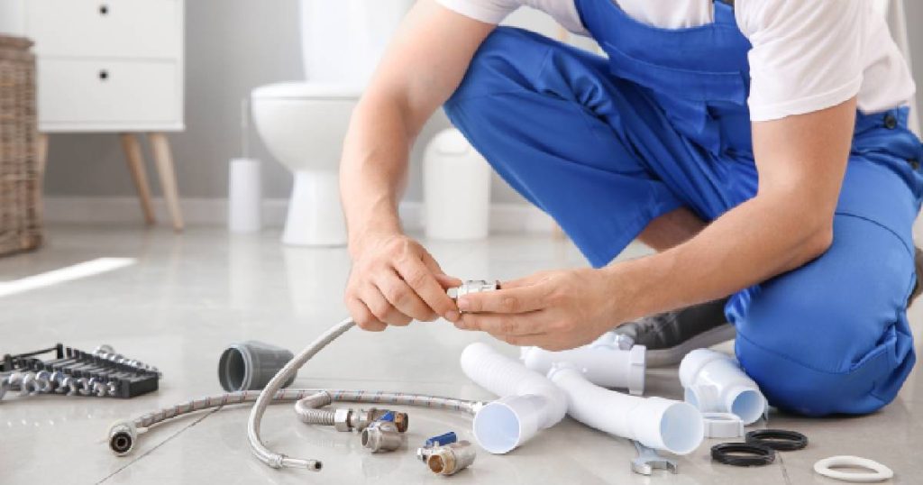 Een man in een wit t-shirt en een blauwe overal installeert een goedkope waterontharder in een badkamer. Voor de knielende man liggen verschillende metalen en plastic koppelstukken. 