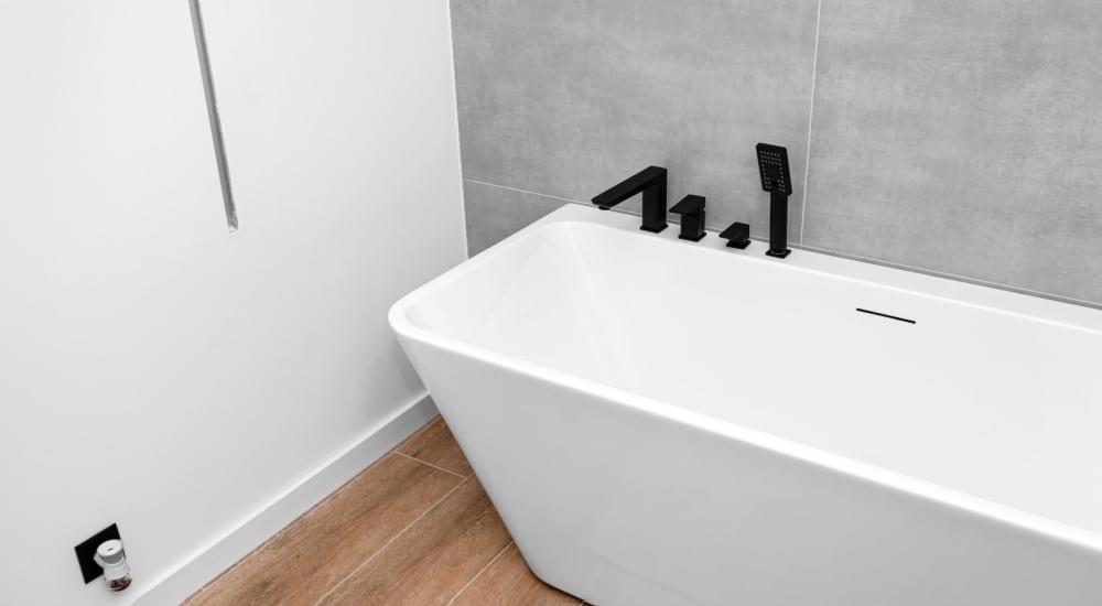 Een wit half vrijstaand bad tegen een grijs betegelde muur met zwarte kranen op de rand.