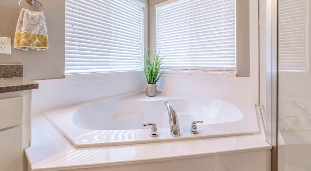 Een ingebouwd wit hoekbad in een witte kleine badkamer net twee ramen, en een wasbak.