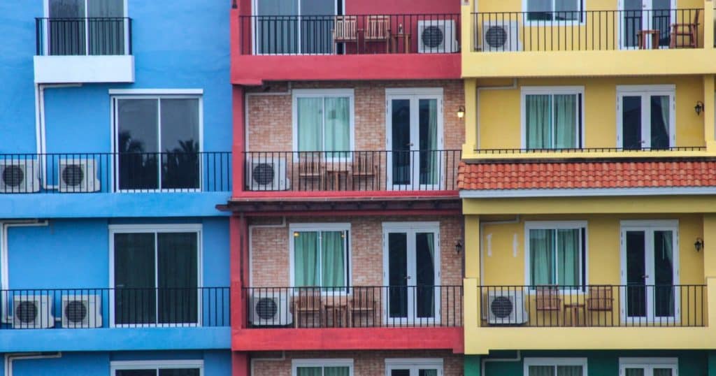Individuele warmtepompen appartementen. Je ziet blauwe, rode en gele appartementen naast elkaar met elk een of meerdere warmtepompen op het balkon.