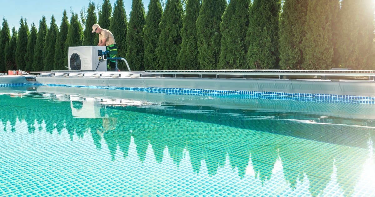 Een specialist installeert een warmtepomp aan een groot zwembad met op de achtergrond een rij groene bomen.