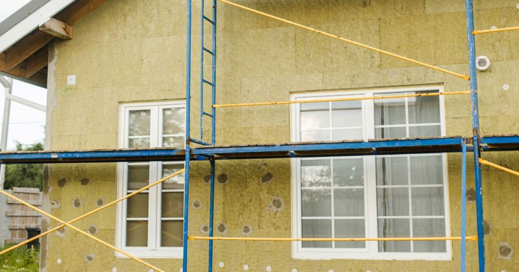Een woning met kale gevels bedekt met houtvezelplaten. Voor de gevel (met twee grote ramen) staat een blauw met gele metalen stelling.