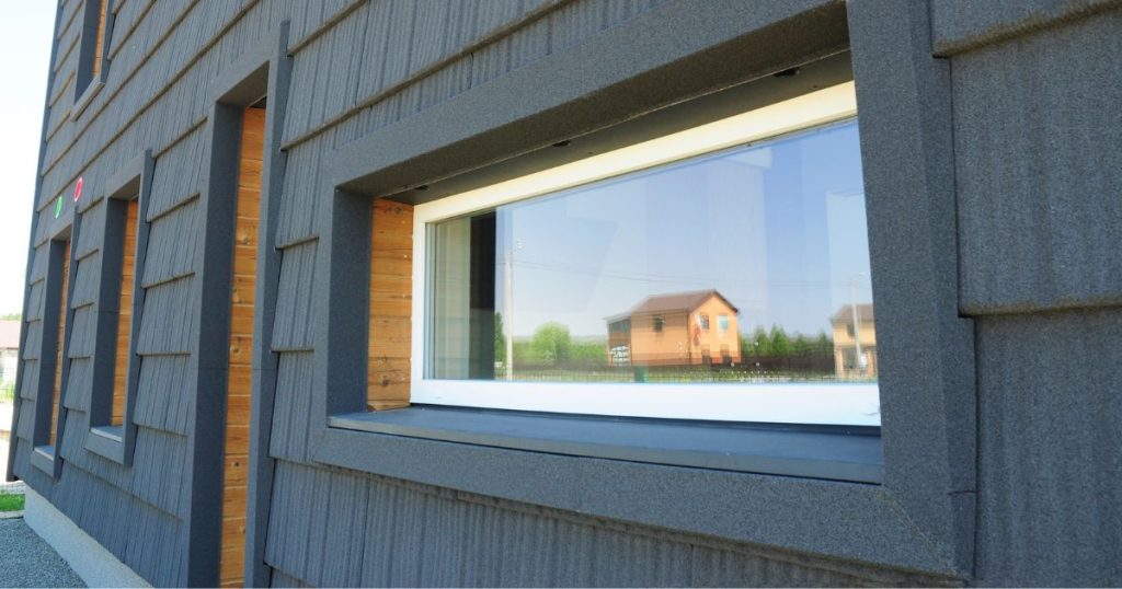 Een raam met witte kozijnen in een gevel met donkergrijze gevelpanelen. Het raam reflecteert een oranje woning in de verte, omgeven door een groen landschap en helderblauwe lucht.
