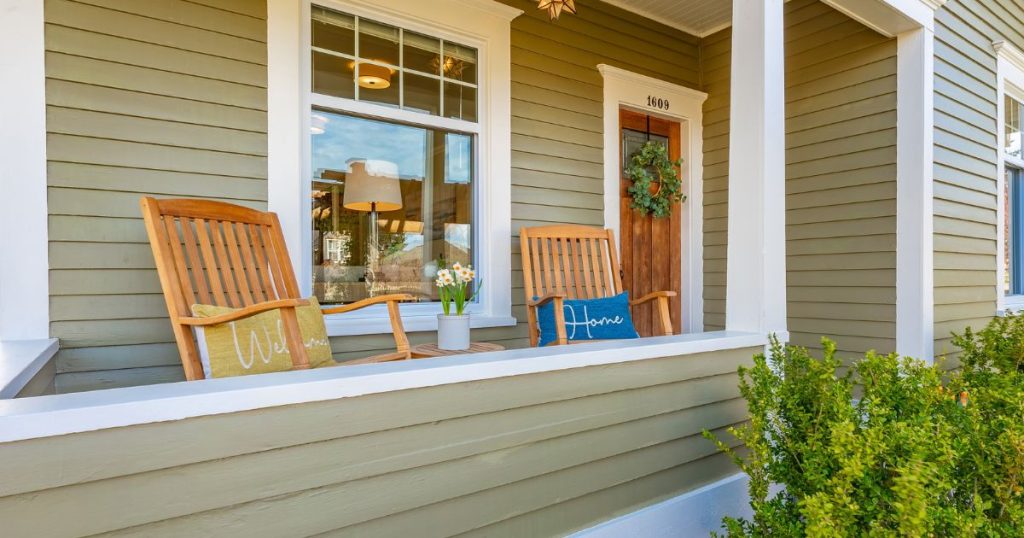 een houten serre aan de voorkant van de woning. de serre is in dezelfde stijl als de woning aangebouwd waardoor het een verlengstuk van de woning is geworden. De serre is open en er staan twee stoelen in, naast de voordeur.