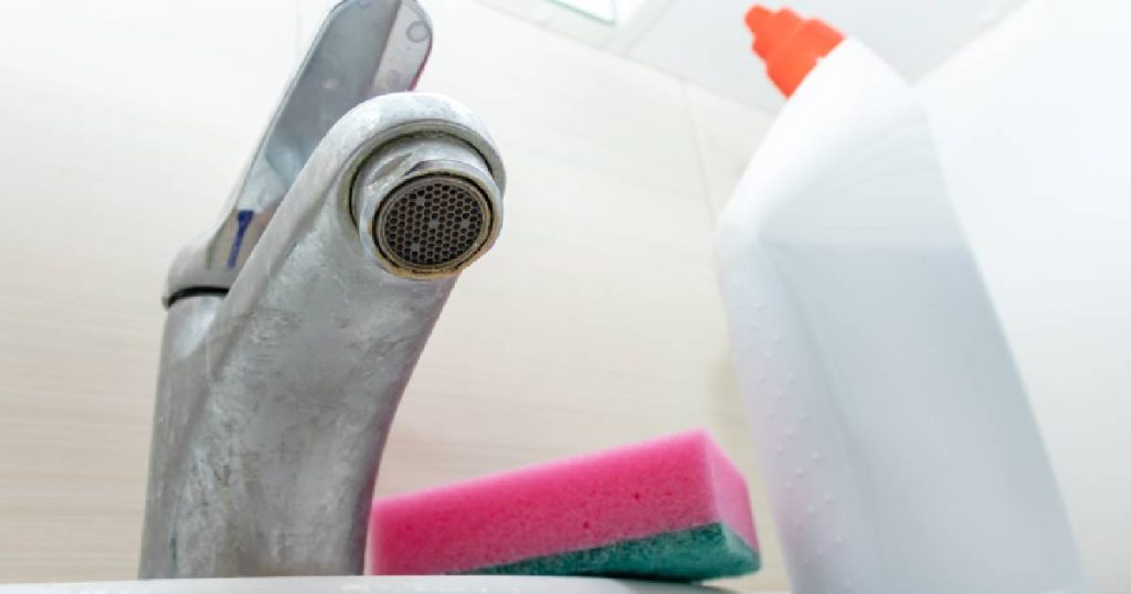 Kikkerperspectief van een kraan met kalkaanslag, een bus schoonmaakazijn en een roze sponsje tegen een witte tegelmuur.