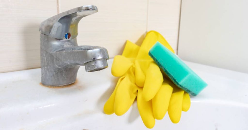 Een kraan met kalkaanslag, twee gele schoonmaakhandschoenen en een groene spons - enkele benodigdheden voor het ontkalken van een kraan.