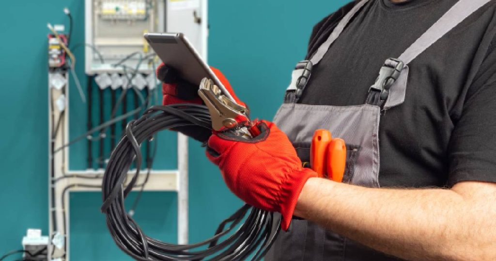 Een elektricien met zwart t-shirt, grijze overal en rode handschoenen inspecteert een 3-fase aansluiting bij een klant thuis waarop een laadpaal is aangesloten. Op de achtergrond is een meterkast zichtbaar, gemonteerd tegen een appelblauwzeegroene muur.