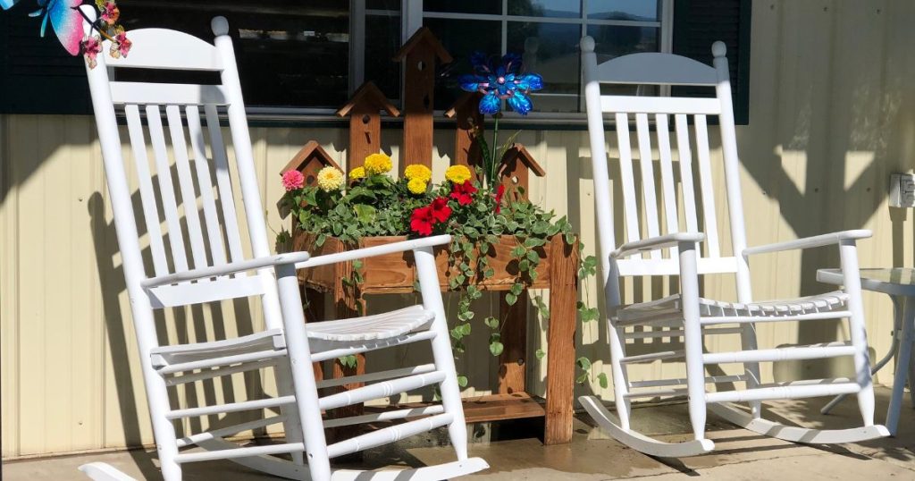 Twee witte houten wipstoelen in een open serre aan de voorkant van een woning. Tussen de twee stoelen is nog een decoratieve houten meubel met planten te zien voor een extra landelijke uitstraling.
