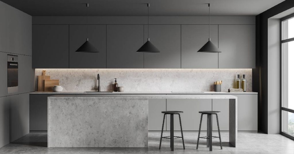 Een moderne, lichtgrijze keuken met minimalistische aankleding. De keuken beschikt over een grijze gietvloer, grijze, betonnen keukeneiland en warmgrijze keukenkasten. Drie donkergrijze hanglampen voegen een vleugje stijl en elegantie toe aan het sobere ontwerp.