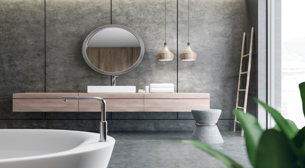 Een wit losstaand bad met een zilveren kraan in een ruime grijze badkamer met zwevende houten kastjes.