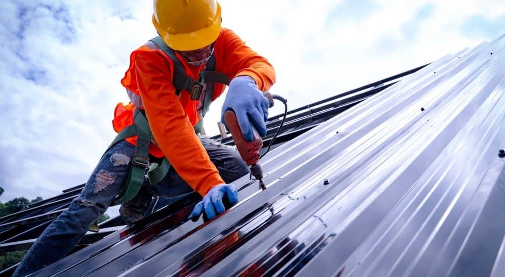 Montage van dakplaten door vakman met oranje trui, gele helm en veiligheidsgordel. De blauwe handschoenen beschermen de handen tegen de ruwe materialen en bij het bedienen van de rode schroefboor.
