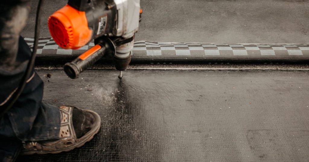 Zwarte dakbedekking op een warm geïsoleerd plat dak. Een dakwerker met werkschoenen gebruikt een zwart, oranje en grijs apparaat om de dakbedekking te bevestigen.