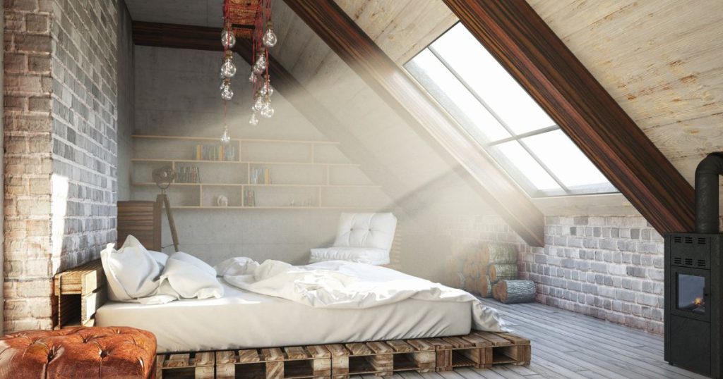 Een slaapkamer onder een hellend dak met een ruim dakraam, palletbed met witte lakens en kussens, een leren poef, zwarte kachel, houtblokken, en wandplanken.