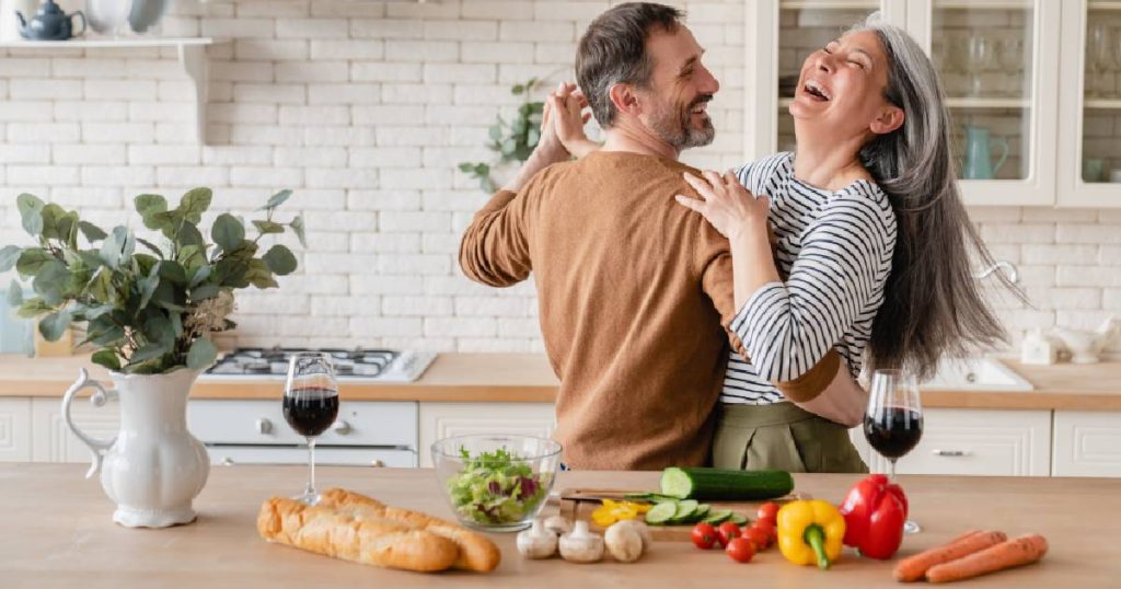 Een koppel danst in hun open parallel keuken met houten aanrecht, plank aan de muur, landelijke tegeltjes en bovenkasten met glazen deurtjes.