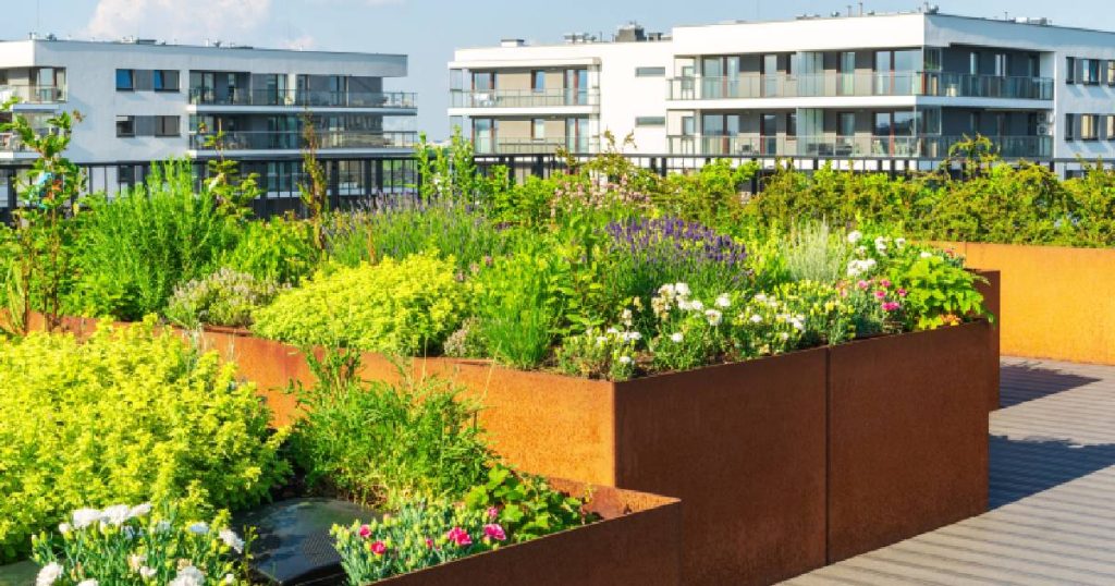 Een huis of appartement met een plat dak met dakterras. Op het dakterras staan roestkleurige plantenbakken met allerlei soorten begroeiing. Het dakterras heeft een houten vloer en heeft een onbelemmerd uitzicht op  de omliggende moderne appartementsgebouwen.