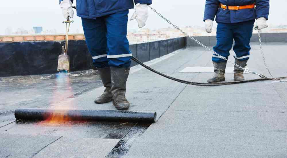 Twee dakspecialisten in blauwe werktenue vernieuwen de roofing op een plat dak. De voorste expert houdt een brander vast om de roofing aan het dak te hechten.