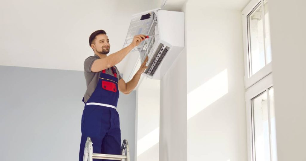 Een installateur in grijs t-shirt en blauw met rode overall installeert een wandmodel Samsung airco in een witte kamer met hoge ramen.